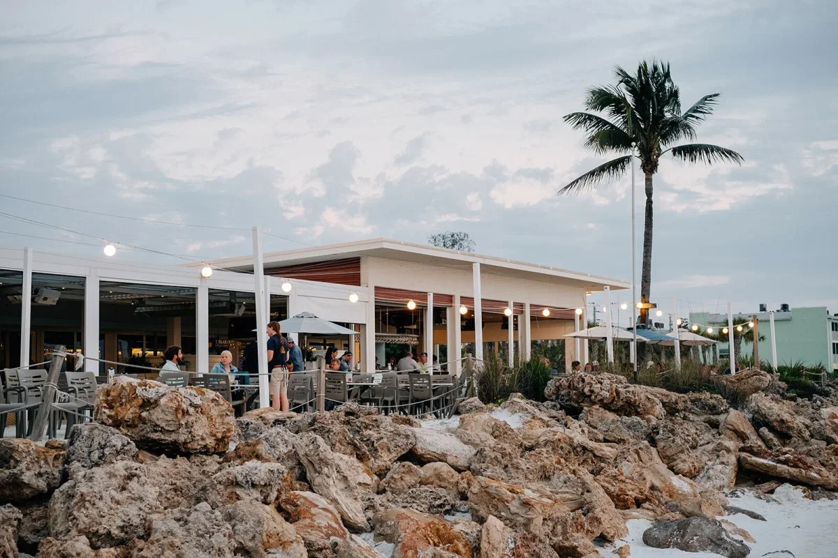 Sarasota Motor Club - Premium Car Service Sarasota - An outdoor seaside restaurant with people dining and staff serving, surrounded by rocks and palm trees, under a partly cloudy sky during sunset or early evening.