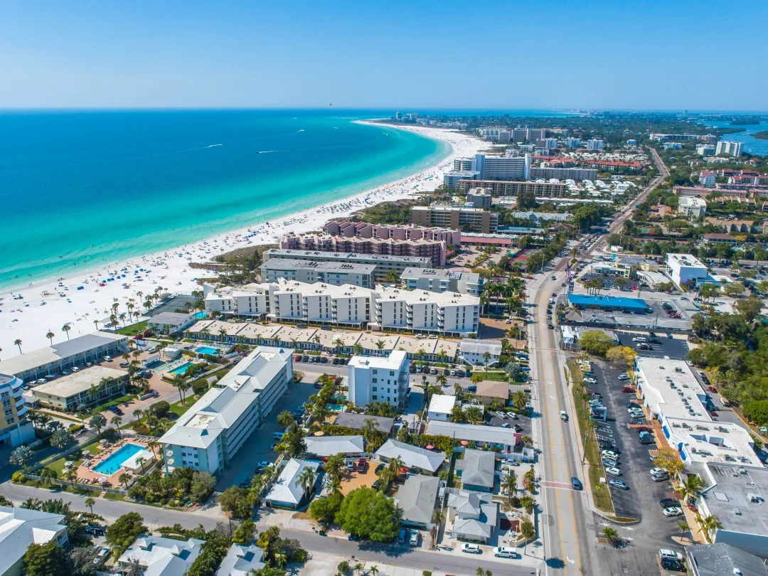 Sarasota Motor Club - Exotic Car Consignment Sarasota - Aerial view of a beachside city with high-rise hotels, condominiums, and a busy street, overlooking a white sandy beach and turquoise ocean.