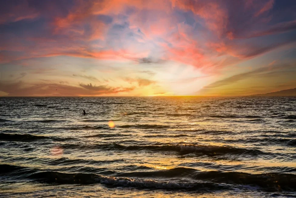 Sarasota Motor Club - Premium Car Service Sarasota - Sunset over the ocean with colorful clouds and gentle waves, with a person in the water.