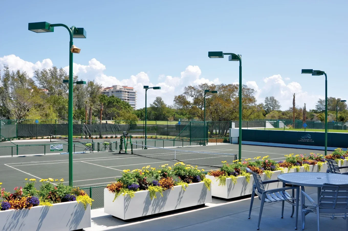 Sarasota Motor Club - Exotic Car Rental Sarasota Florida - Outdoor tennis courts with green fencing, surrounded by colorful flower planters and metal outdoor furniture, under a partly cloudy sky.