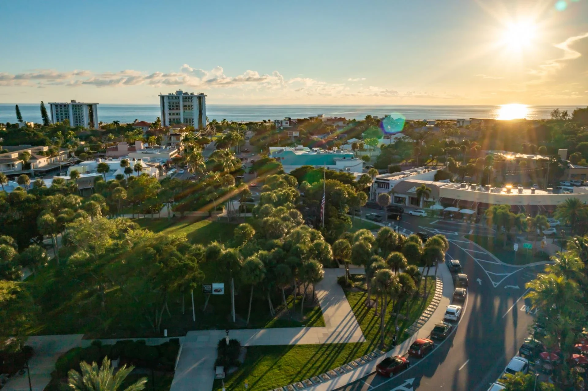 Sarasota Motor Club - Exotic Car Consignment Sarasota - Aerial view of a city near the coast at sunset, with trees, buildings, and streets, and the ocean in the background.