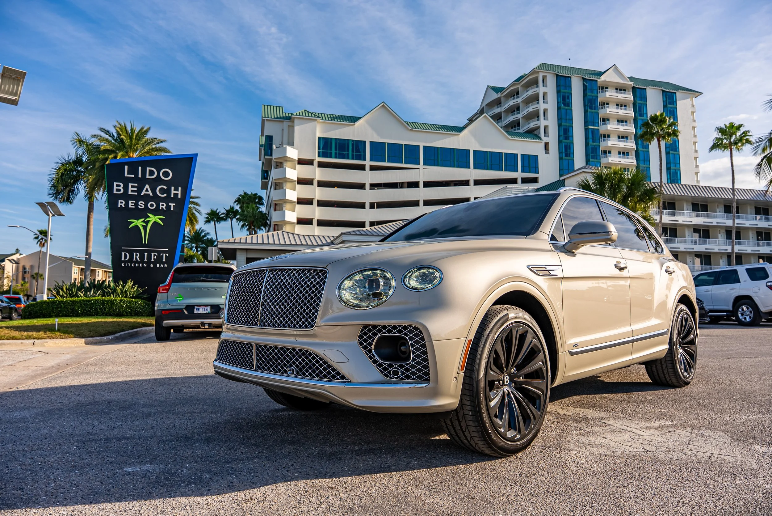 Sarasota Motor Club - Lido key exotic car rental - A cream-colored Bentley parked in front of the Lido Beach Resort with a large building, palm trees, and a clear blue sky in the background.