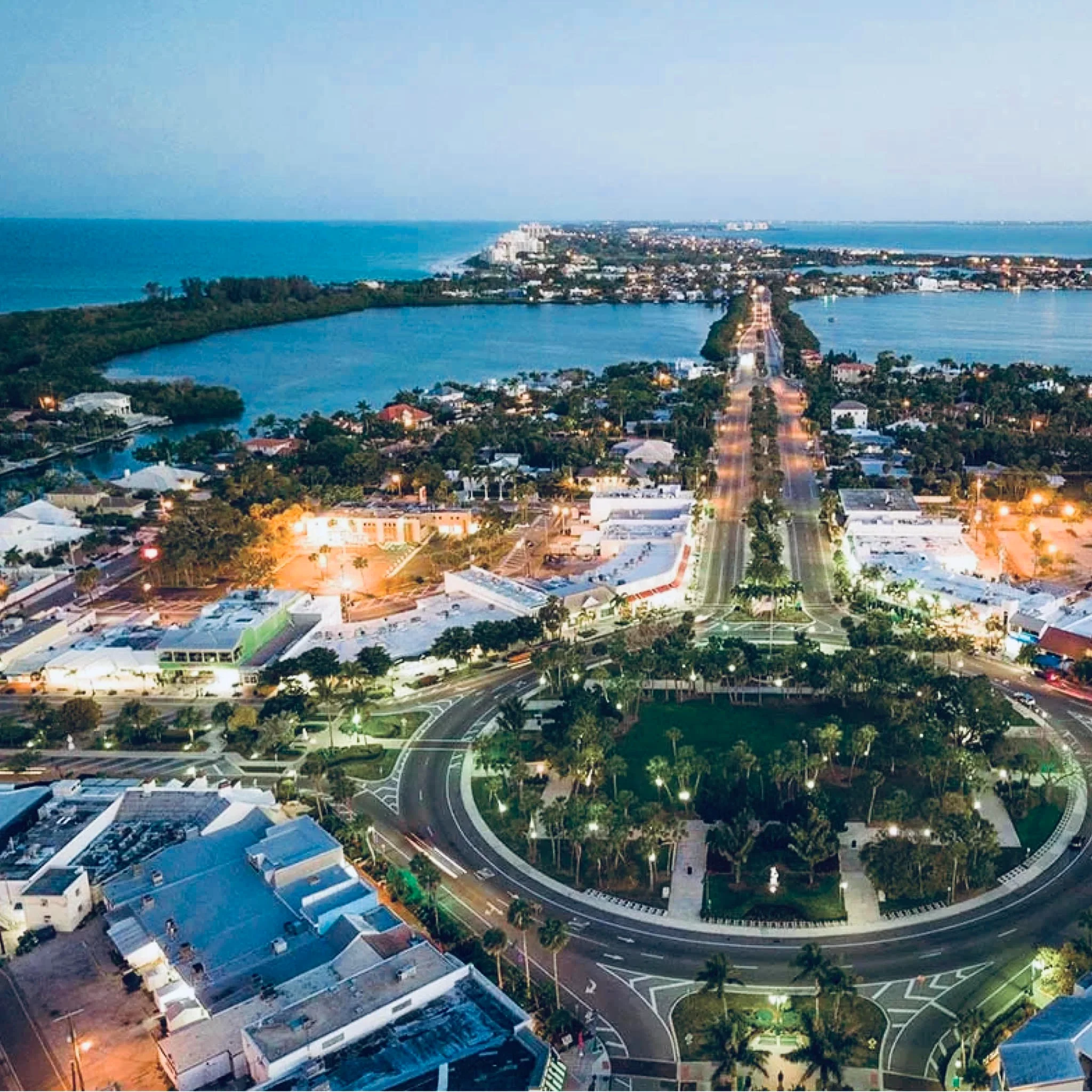 Sarasota Motor Club - Sarasota Luxury Car Rental - Aerial view of a coastal town with a circular park and a waterway in the background during evening.
