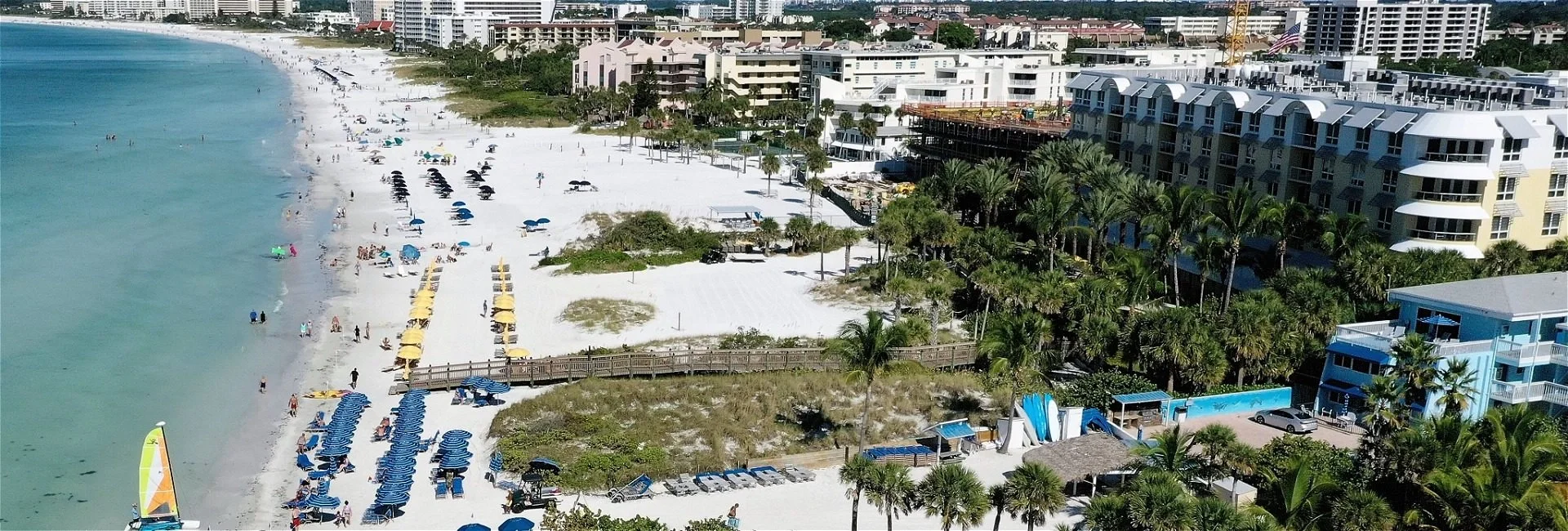 Sarasota Motor Club - Premium Car Service Sarasota - Aerial view of a beach with white sand, blue water, thatched umbrellas, and lounge chairs, with buildings and palm trees in the background.