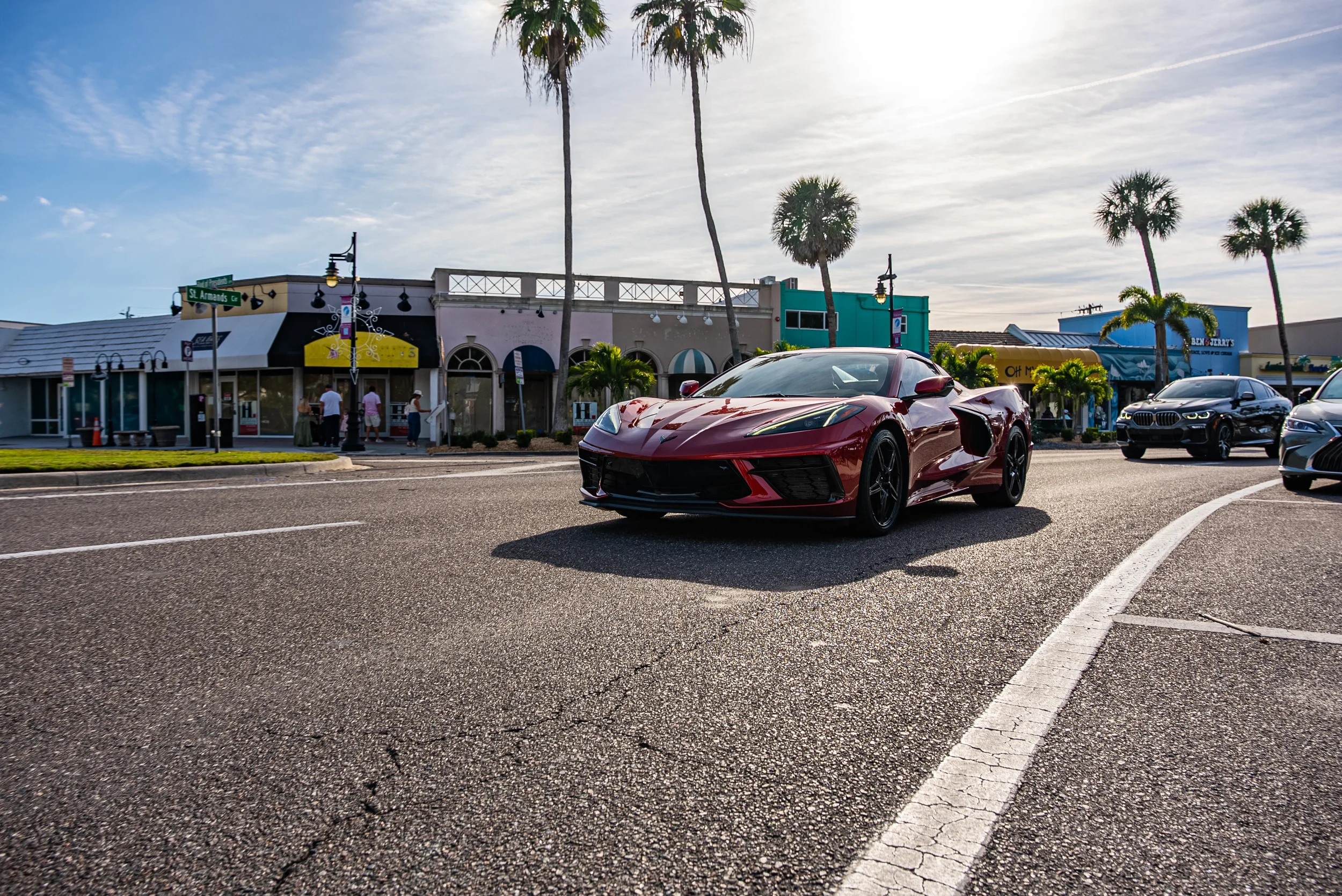 Sarasota Motor Club - premium car service sarasota - A red sports car driving on a city street lined with palm trees, shops and pedestrians under a partly cloudy sky.