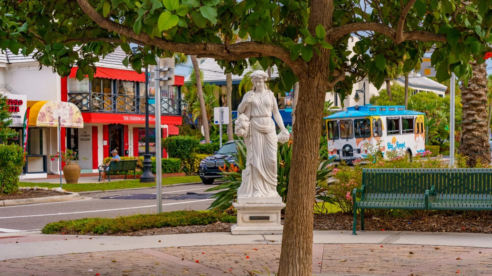Sarasota Motor CA small city park with a marble statue of a woman holding flowers, a tree in the foreground, a green park bench, and a colorful trolley bus with pink and blue decorations. Behind are shops with awnings and a person sitting on a bench.