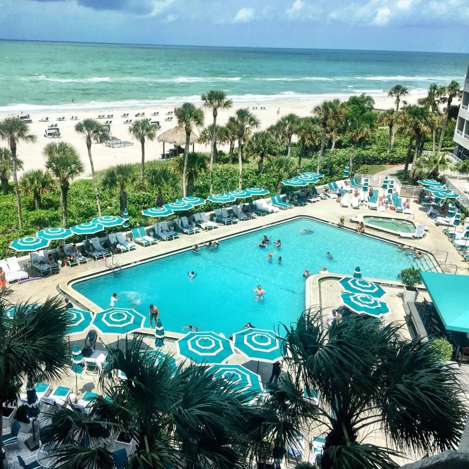 Sarasota Motor Club - Sarasota Luxury Car Rental - Swimming pool with people swimming and lounging around, surrounded by blue and white umbrellas, with a beach and ocean in the background.