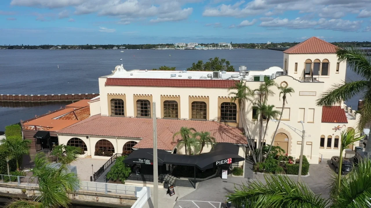 Sarasota Motor Club - Exotic Car Rental Sarasota Florida - A white Mediterranean-style building with a red-tiled roof, arched windows, and a rooftop balcony, situated along a waterfront with palm trees and a marina in the background.
