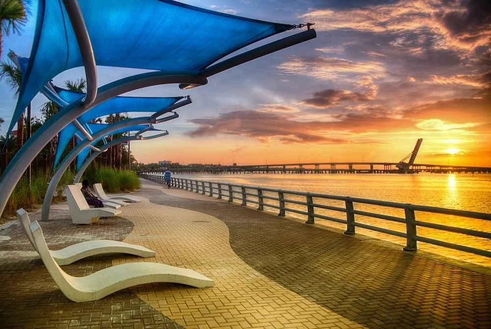 Sarasota Motor Club - Exotic Car Rental Sarasota Florida - Sunset over a riverside walkway with modern white benches and blue shade structures, a person walking, and a bridge in the background.