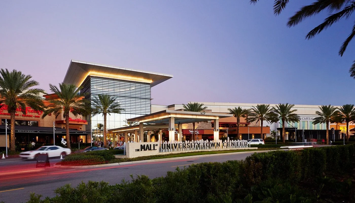 Sarasota Motor Club - Exotic Car Rental Sarasota Florida - Exterior view of Maitland University Town Center shopping mall with palm trees, parked cars, and stores during dusk.