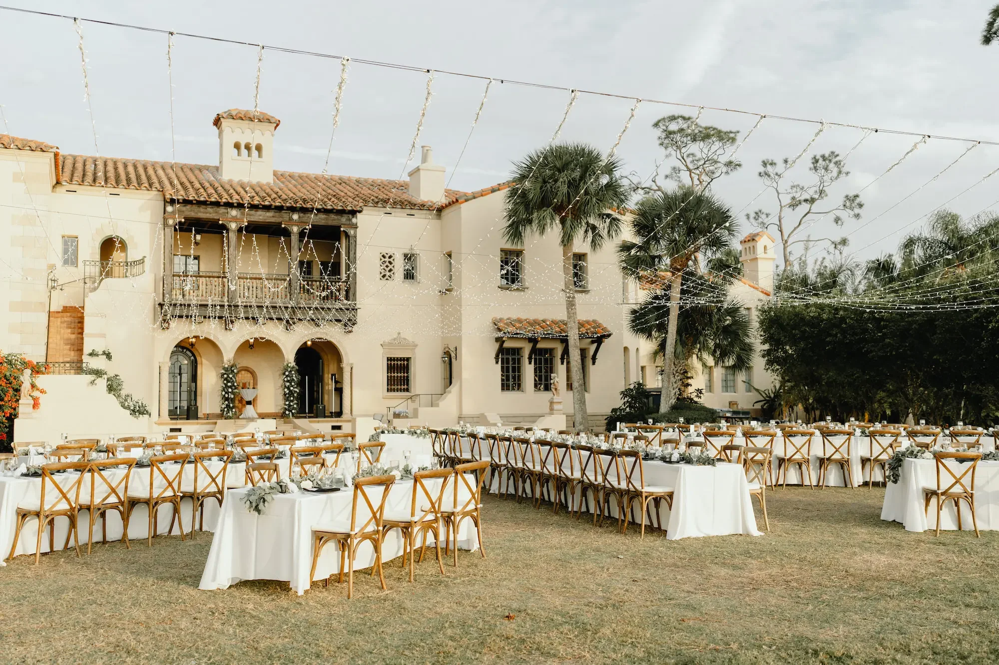 Outdoor wedding reception setup with long tables covered in white tablecloths, decorated with floral arrangements, set with plates, glasses, and silverware, in front of a large mansion with Mediterranean architecture, palm trees, and string lights hanging overhead.