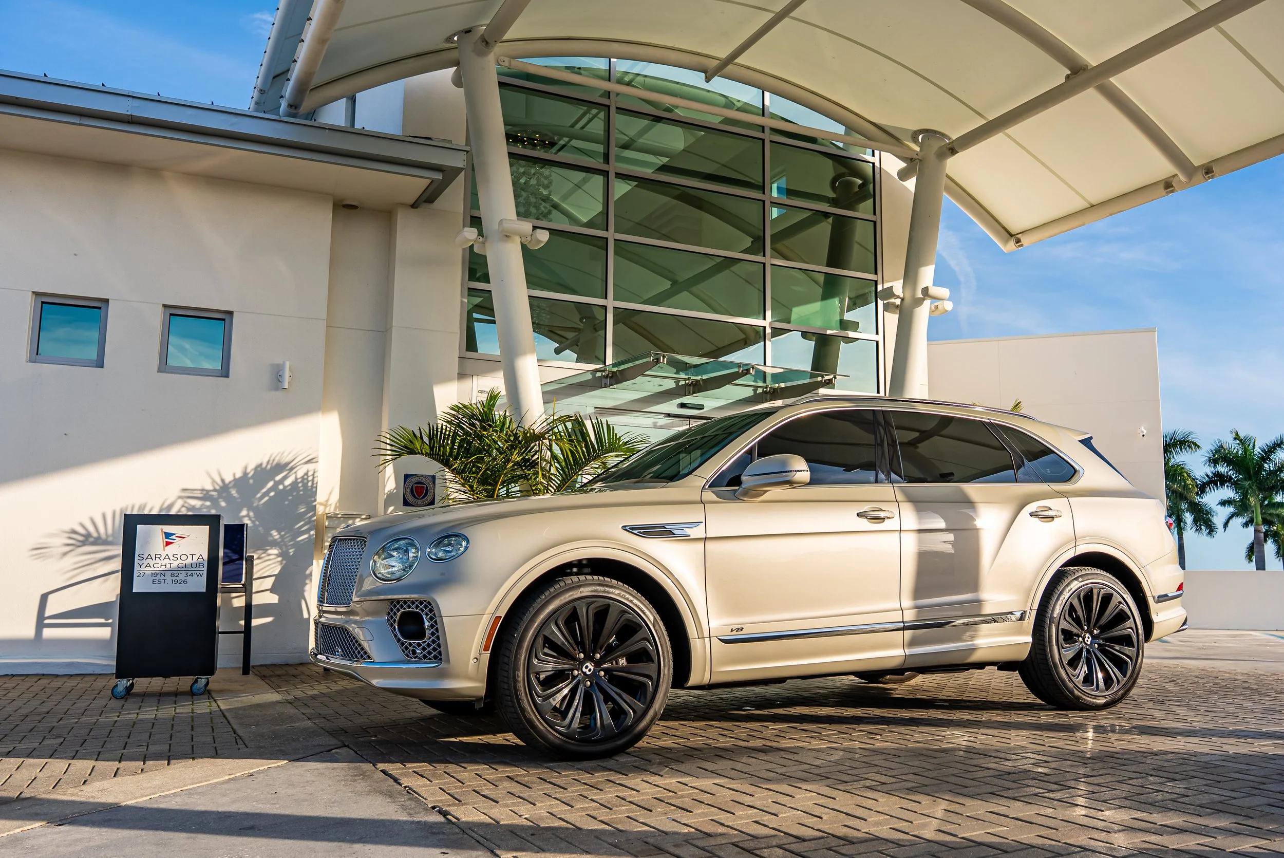 Sarasota Motor Club -  luxury car rental sarasota - A beige Bentley Bentayga parked in front of the Sarasota Yacht Club with palm trees in the background.