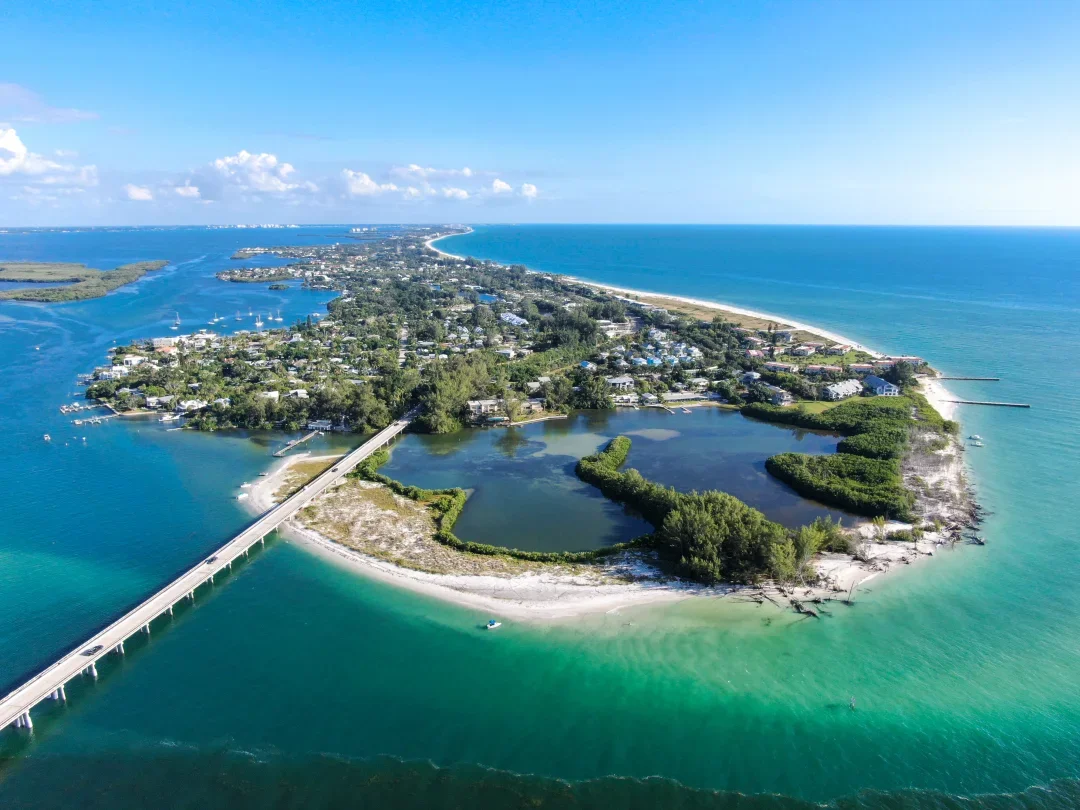 Sarasota Motor Club - Sports Car Rental Sarasota FL - Aerial view of a coastal town with a bridge connecting to a peninsula surrounded by water and lush greenery, with the ocean in the background.