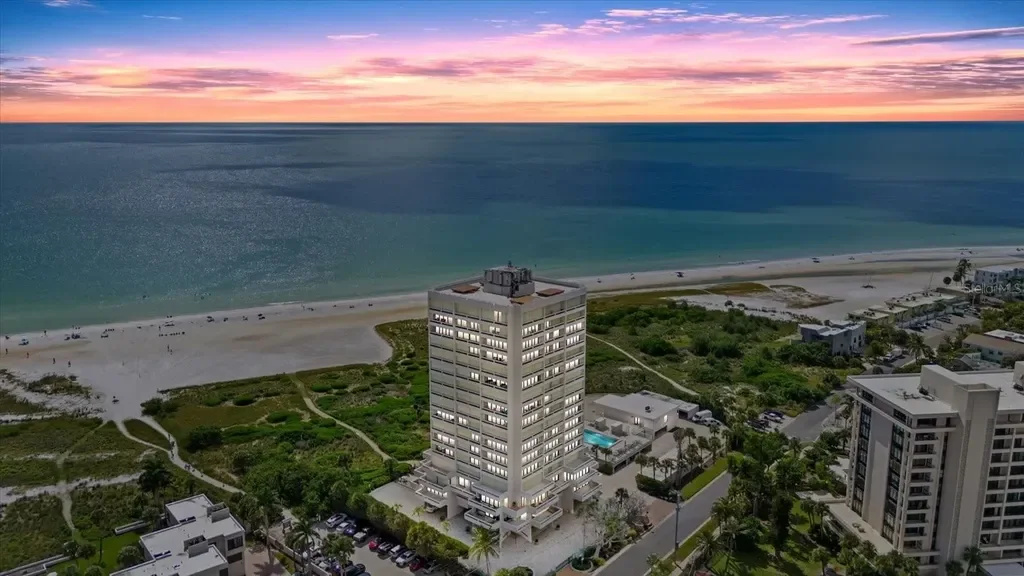 Sarasota Motor Club - Sports Car Rental Sarasota FL - Aerial view of a beachfront hotel with a swimming pool, surrounded by other buildings, overlooking a sandy beach and ocean at sunset.