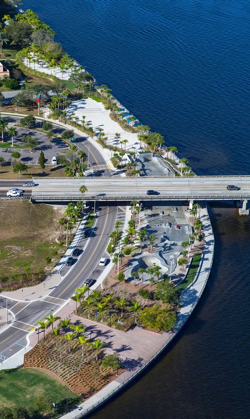 Sarasota Motor Club - Exotic Car Consignment Sarasota - Aerial view of a waterfront park featuring palm trees, a sandy beach area, pathways, parking lot, and a bridge over water.