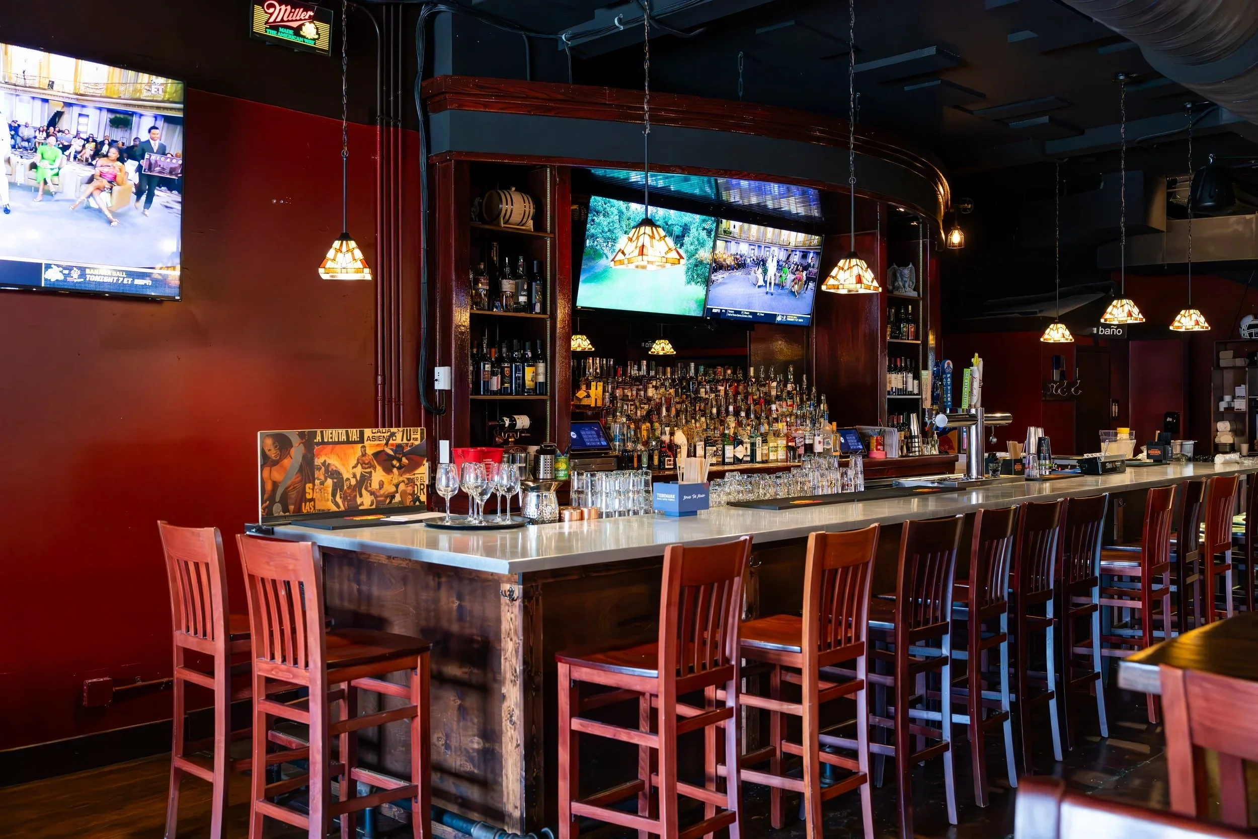 Sarasota Motor Club - Premium Car Service Sarasota - Interior of a bar with a white marble countertop, bar stools, shelves with various bottles of alcohol, television screens, and hanging lights.