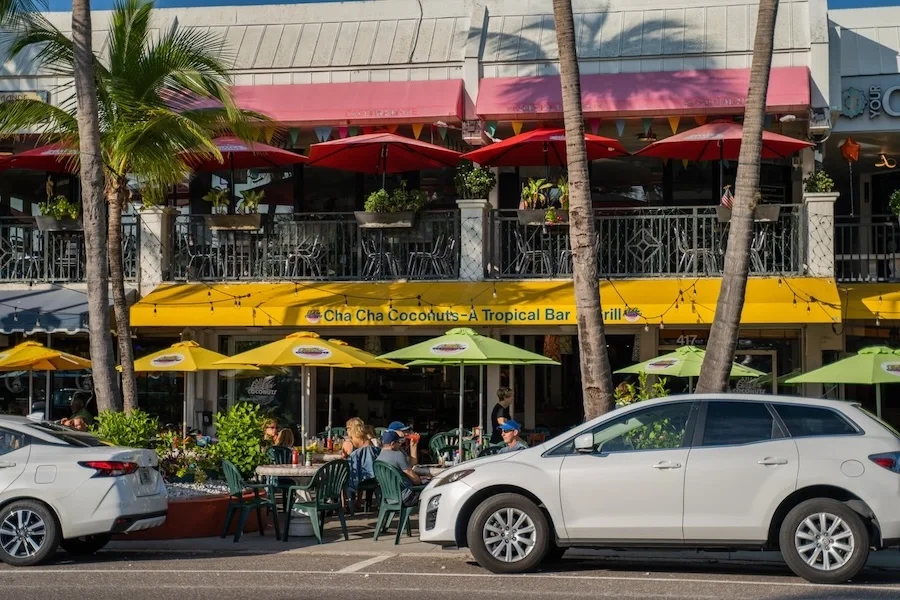 Sarasota Motor Club - Exotic Car - Street scene with outdoor seating at a restaurant called "Cha Cha Coconuts - A Tropical Bar & Grill" with yellow and green umbrellas, palm trees, and people dining. Two cars parked in front of the restaurant.
