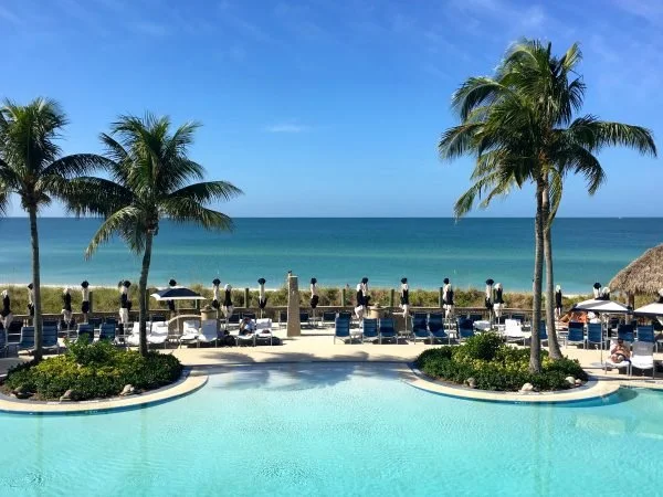 Sarasota Motor Club - casey key luxury car rental - Luxury poolside view with palm trees, lounge chairs, and umbrellas overlooking the ocean on a sunny day.