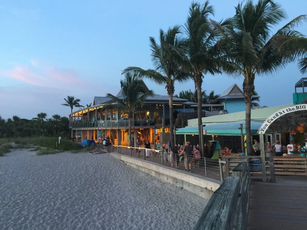 Sarasota Motor Club - Premium Car Service Sarasota - Beachfront restaurant with palm trees and outdoor seating, people dining and socializing during early evening.