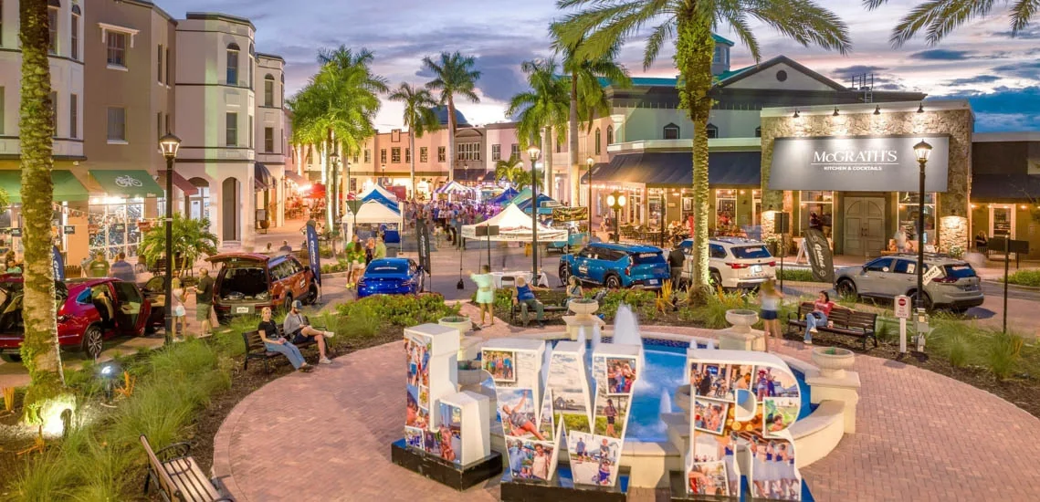 A lively shopping and dining district at dusk with palm trees, parked cars, outdoor seating, tents, and people walking. There is a fountain with large letters spelling 'LIVR' decorated with photos in the foreground and streetlights illuminating the area.