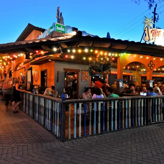 Sarasota Motor Club - Premium Car Service Sarasota - People dining outside at a restaurant with string lights, a wooden railing, and outdoor seating during dusk.