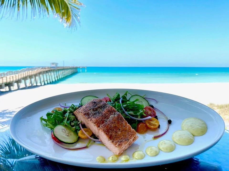 Sarasota Motor Club - Premium Car Service Sarasota - Plate of grilled salmon with fresh salad and sauce, set against a beach and pier background.