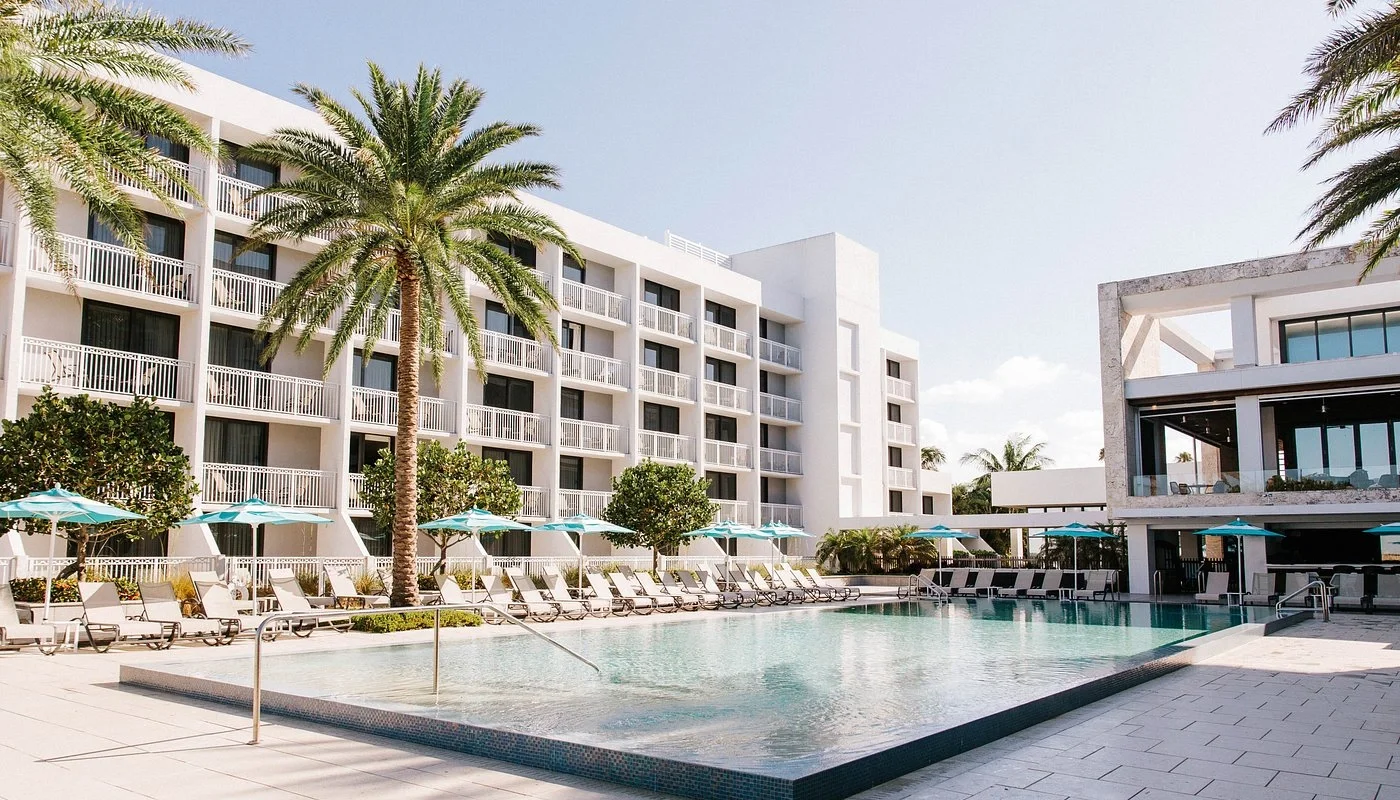 Sarasota Motor Club - Exotic Car Rental Sarasota Florida - A modern hotel pool area with lounge chairs and umbrellas, surrounded by palm trees and white buildings under a clear sky.