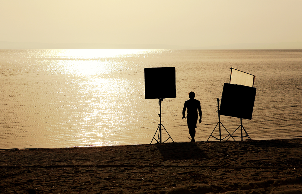Sarasota Motor Club - Exotic Car Rental Sarasota Florida - A silhouette of a person walking on a beach at sunset or sunrise, with photography equipment on stands nearby, facing the water.