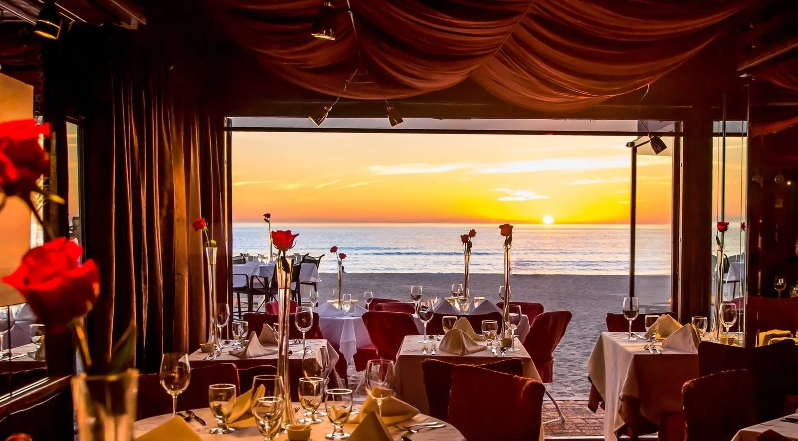 Sarasota Motor Club - Exotic Car Rental Sarasota Florida - A beachside restaurant with tables set for dining, overlooking the ocean at sunset, with flowers and wine glasses on the tables.