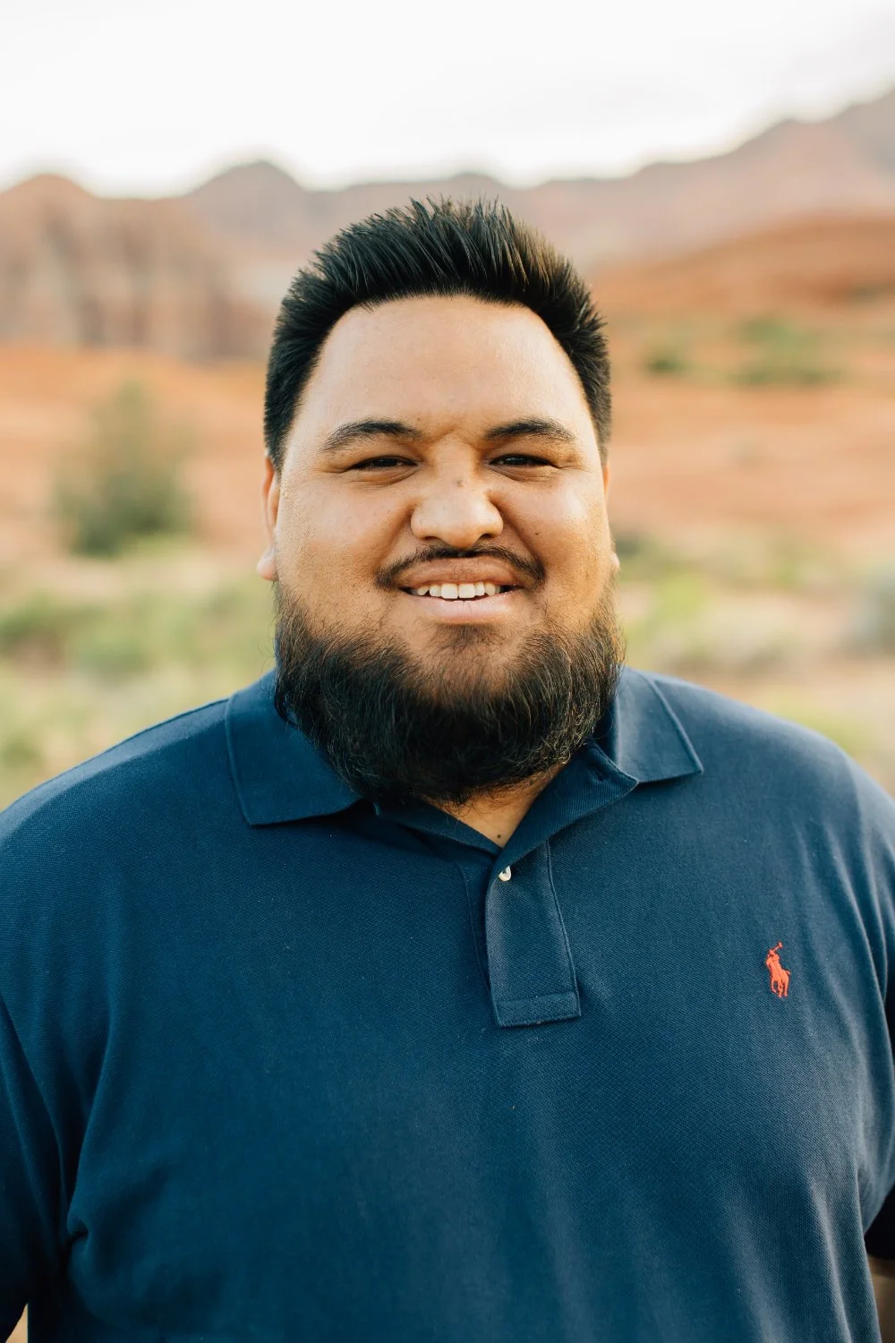 A man with black hair and a beard smiling outdoors with a desert landscape and mountains in the background.