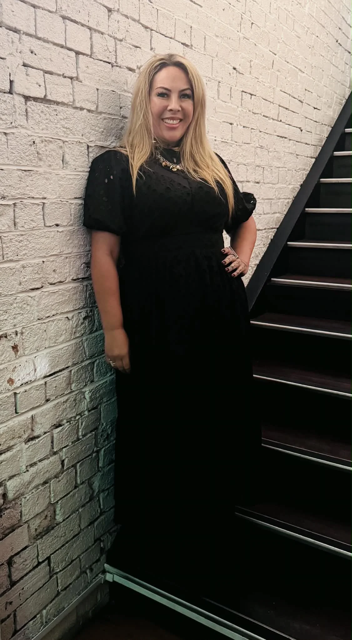 A woman, Helenā Kaho, has blonde hair, wearing a black dress and a gold necklace, poses next to a brick wall and a staircase.