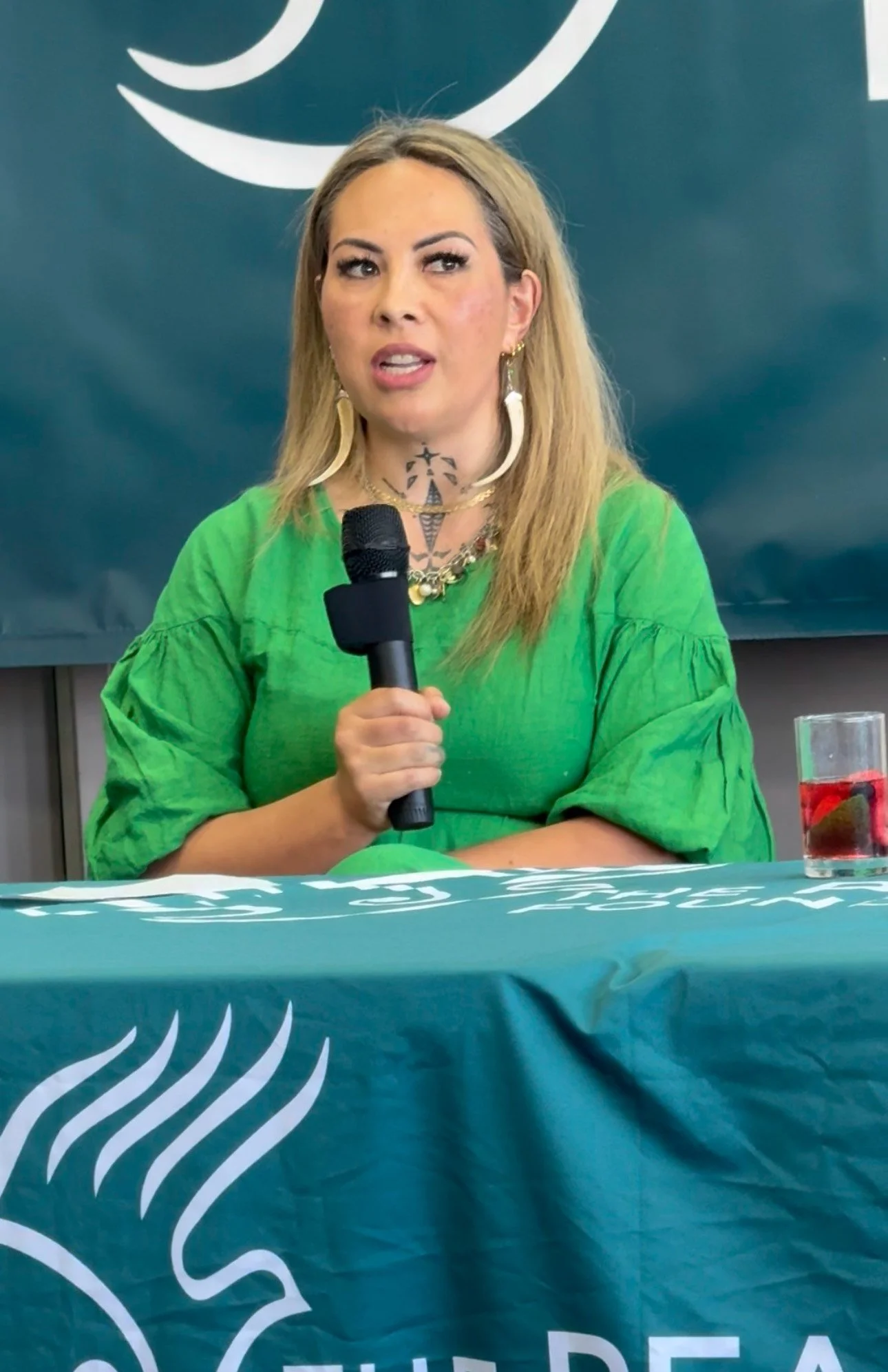 A woman with long blonde hair, wearing a green shirt, holding a microphone, sitting at a table with a blue tablecloth and a glass of red drink. She is speaking at a press conference or event.