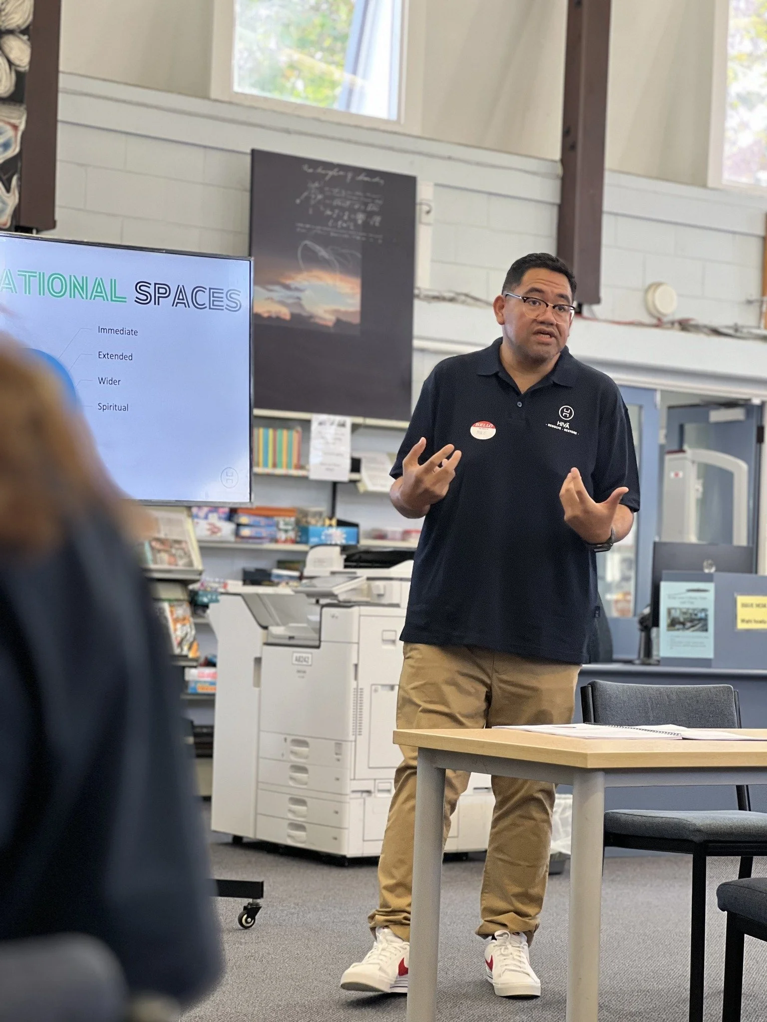 A man giving a presentation in a classroom or conference room. He is wearing glasses, a dark polo shirt, khaki pants, and white sneakers with red accents. There is a large screen behind him displaying a slide titled 'NATIONAL SPACES' with bullet points. The room has white brick walls, large windows, and office equipment such as a copier and shelves with books and supplies.