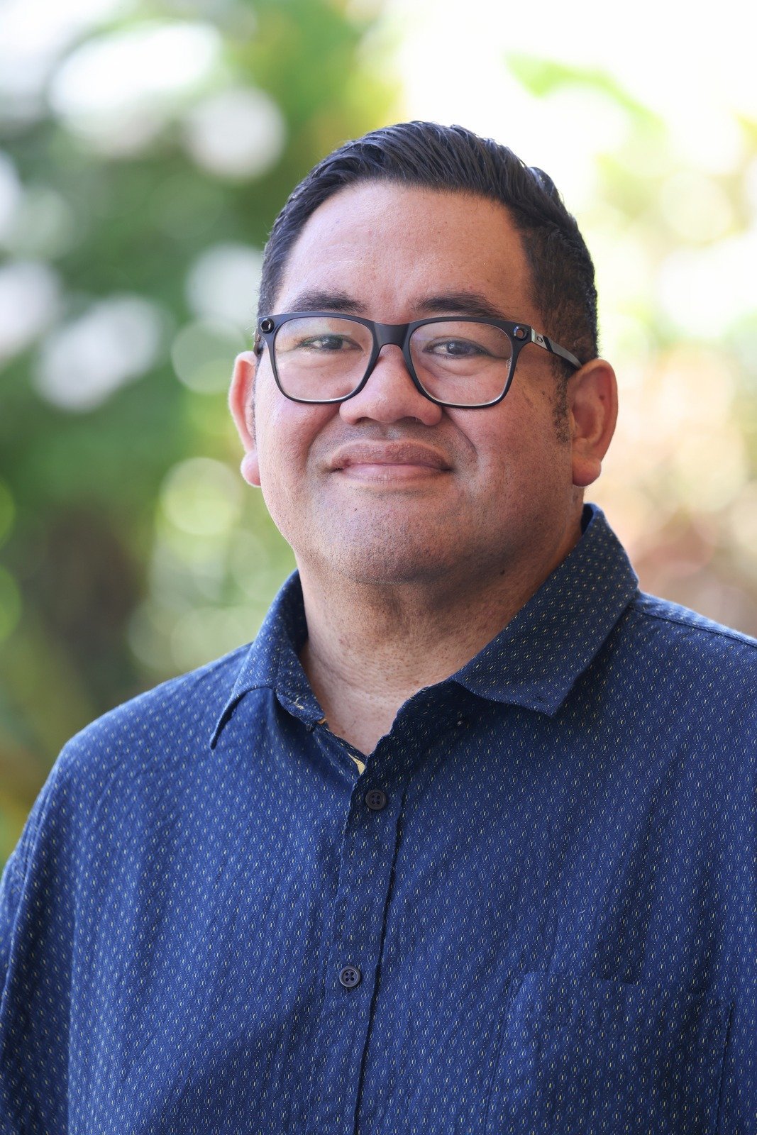 Portrait of a man wearing glasses and a blue patterned shirt, outdoors with a blurred green and yellow background.