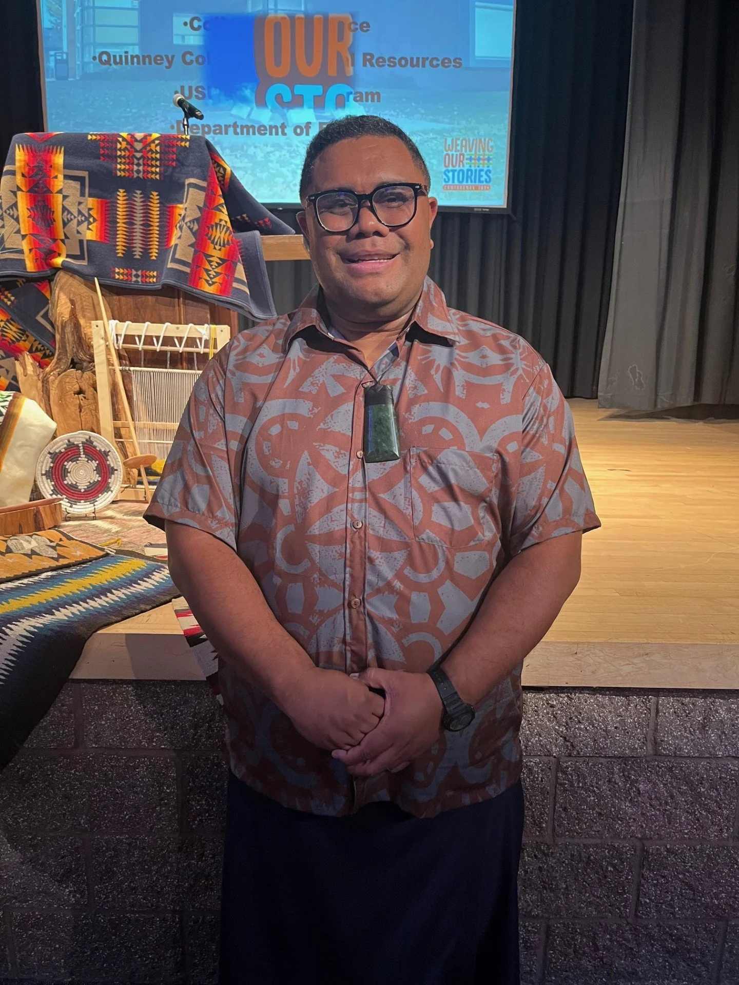A man standing in front of a stage with a large screen displaying the text 'Our Stories'. He is wearing glasses, a patterned shirt, and a black skirt with a watch on his left wrist. The stage behind him has traditional woven items, a blanket with geometric patterns, and a microphone. The background features dark curtains and a wood floor.