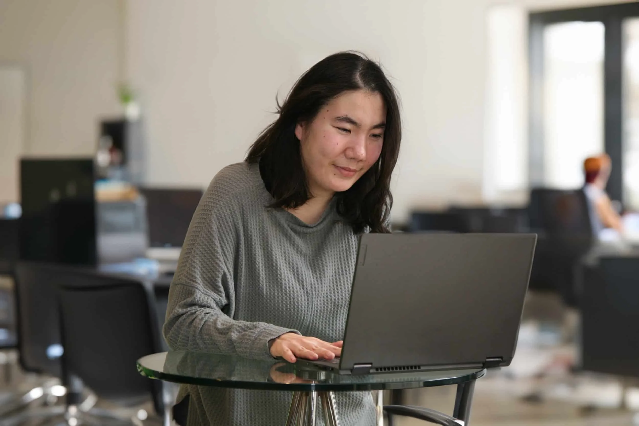 A woman with shoulder-length dark hair working on a laptop at a glass table in an office, with other desks and people in the background.