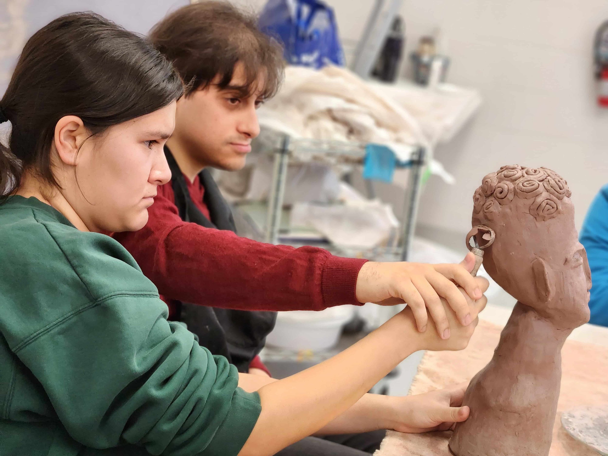Two students working on a clay sculpture of a head with curly hair in an art classroom.