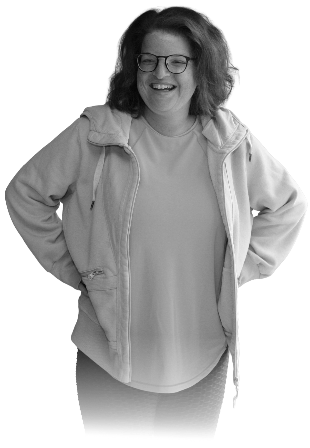 A woman with glasses smiling and standing with hands on hips, wearing a light-colored hoodie and dark patterned leggings.