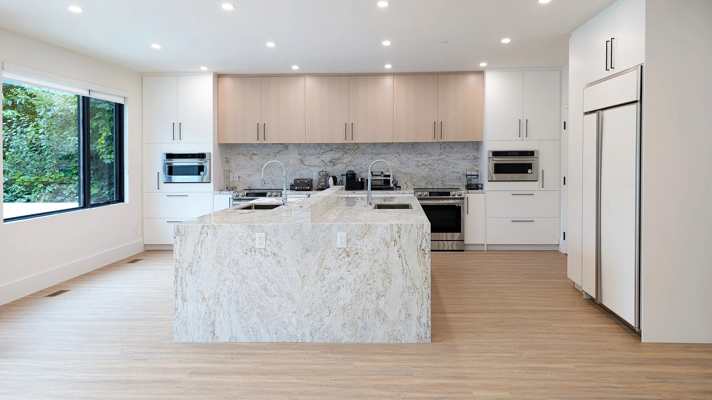 Modern kitchen featuring a large marble island with two sinks, white cabinetry, stainless steel appliances, a marble backsplash, and a large window overlooking greenery.