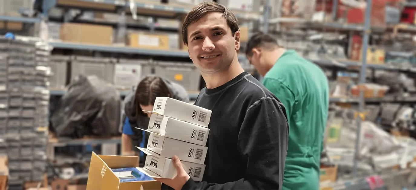 A young man smiling at the camera holding a box in a warehouse with shelves filled with various goods and other people working.