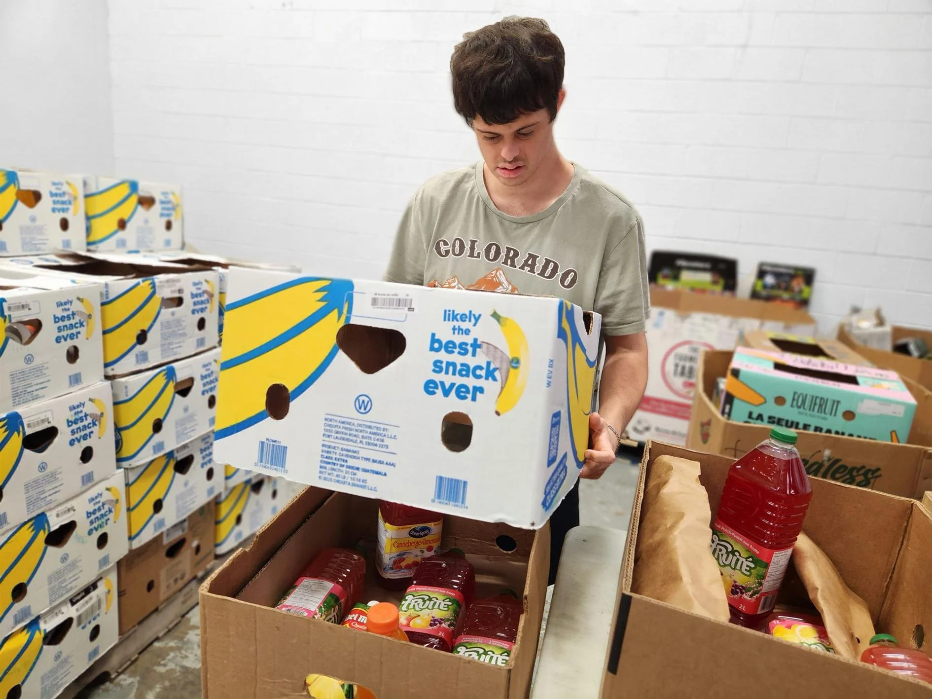 A young man with dark hair wearing a gray T-shirt with 'Colorado' written on it, standing in a warehouse or store aisle, holding a box of bananas labeled 'likely the best snack ever,' surrounded by boxes of grocery items including juice bottles and cardboard crates.