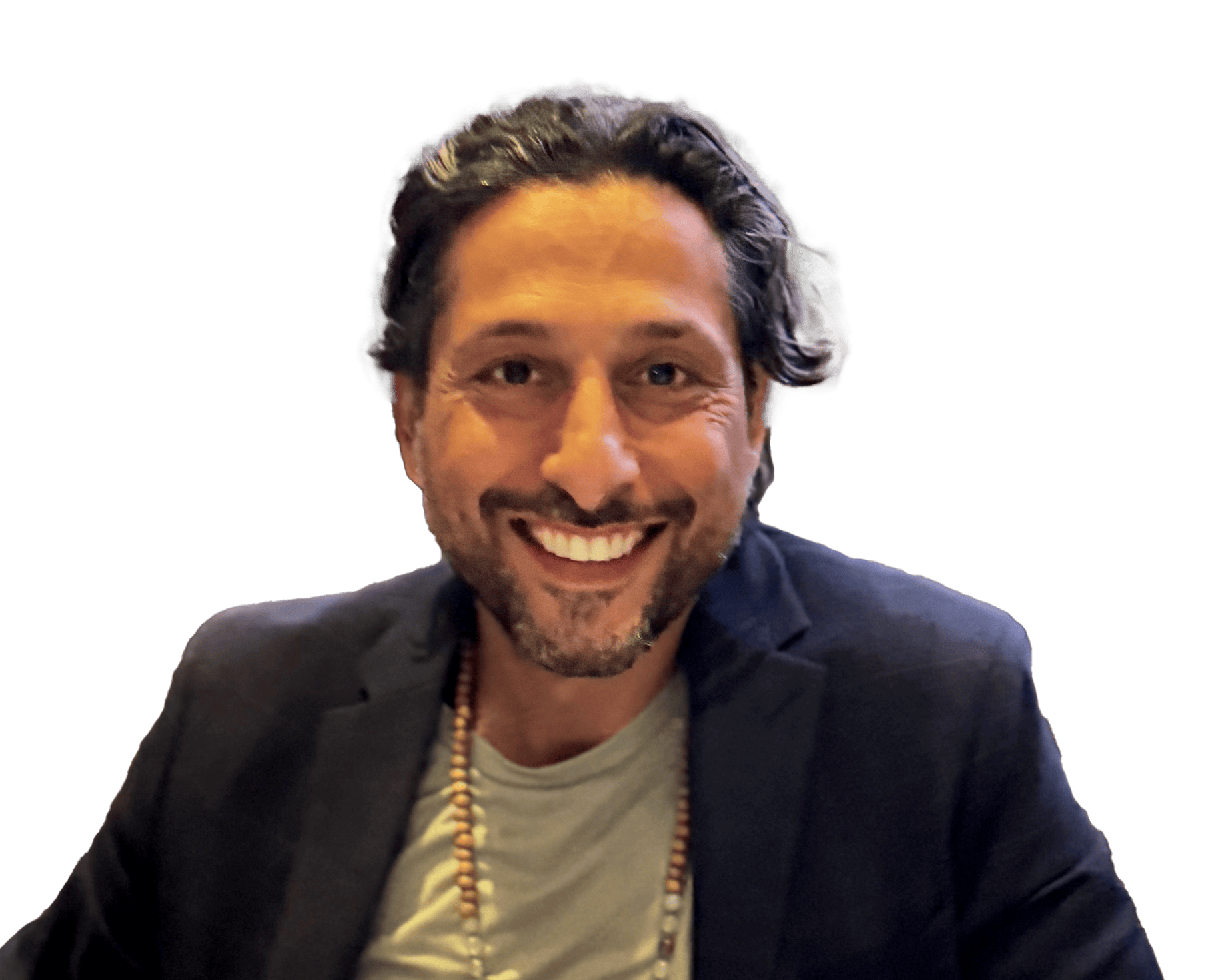 A smiling man with dark hair and a beard, wearing a black blazer, gray T-shirt, and a beaded necklace, sitting in front of a black background.