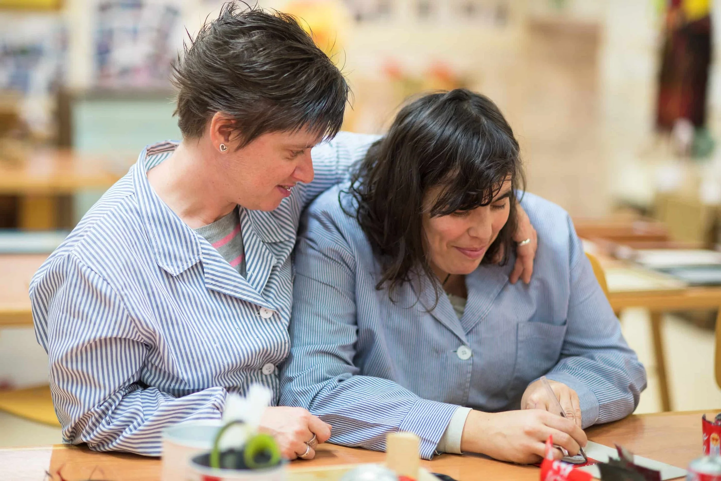 Two women wearing blue striped shirts are sitting closely together at a table, working on arts and crafts with a marker or pen, with one woman resting her chin on the other's shoulder and both smiling.