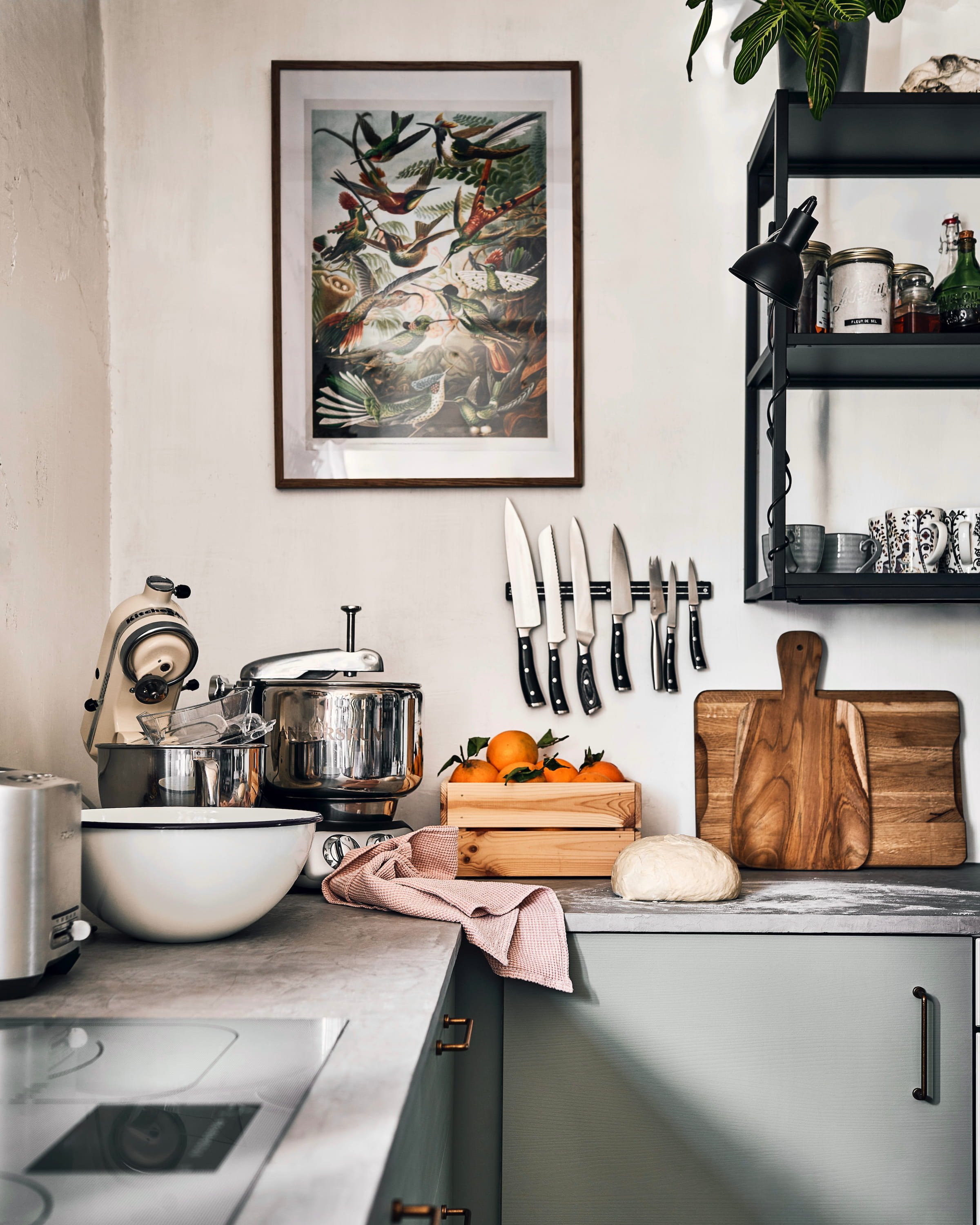 Roheliste köögikappidega taluköök, seinal noad, pilt lindudega ning kappidel lõikelauad, mikserid ja kast apelsinidega