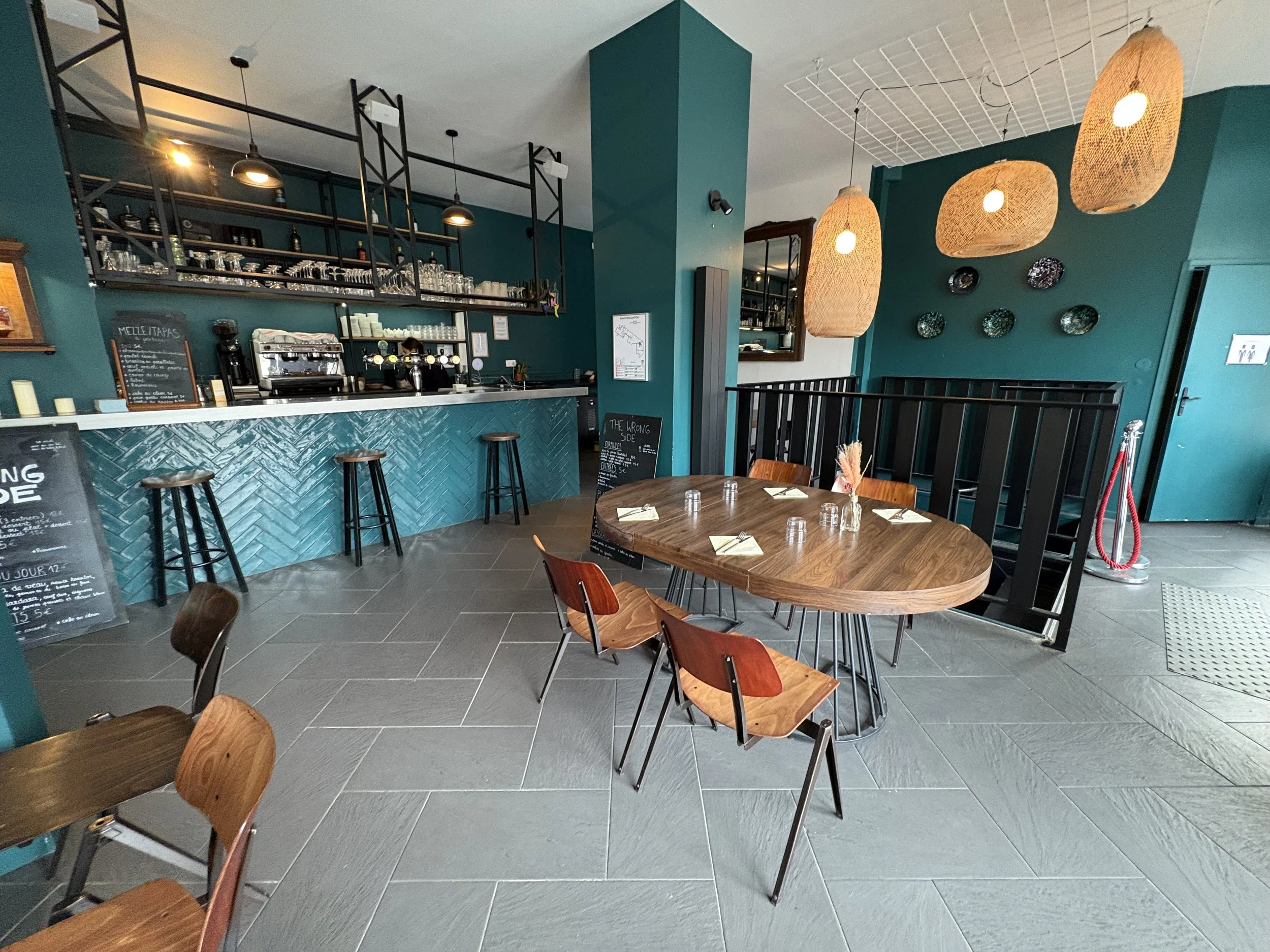 Intérieur d'un café avec un comptoir de bar turquoise, tables en bois, chaises en bois, décoration moderne, grands luminaires en osier suspendus.