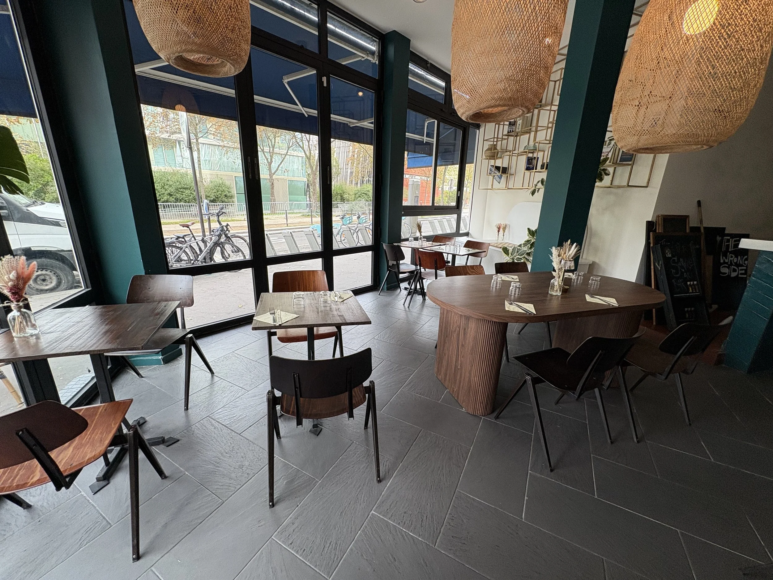 Intérieur d'un café avec des tables en bois, des chaises en bois et en plastique, des lampes tressées suspendues au plafond, des fenêtres grandes laissant entrer la lumière naturelle, et un décor moderne avec des étagères murales.