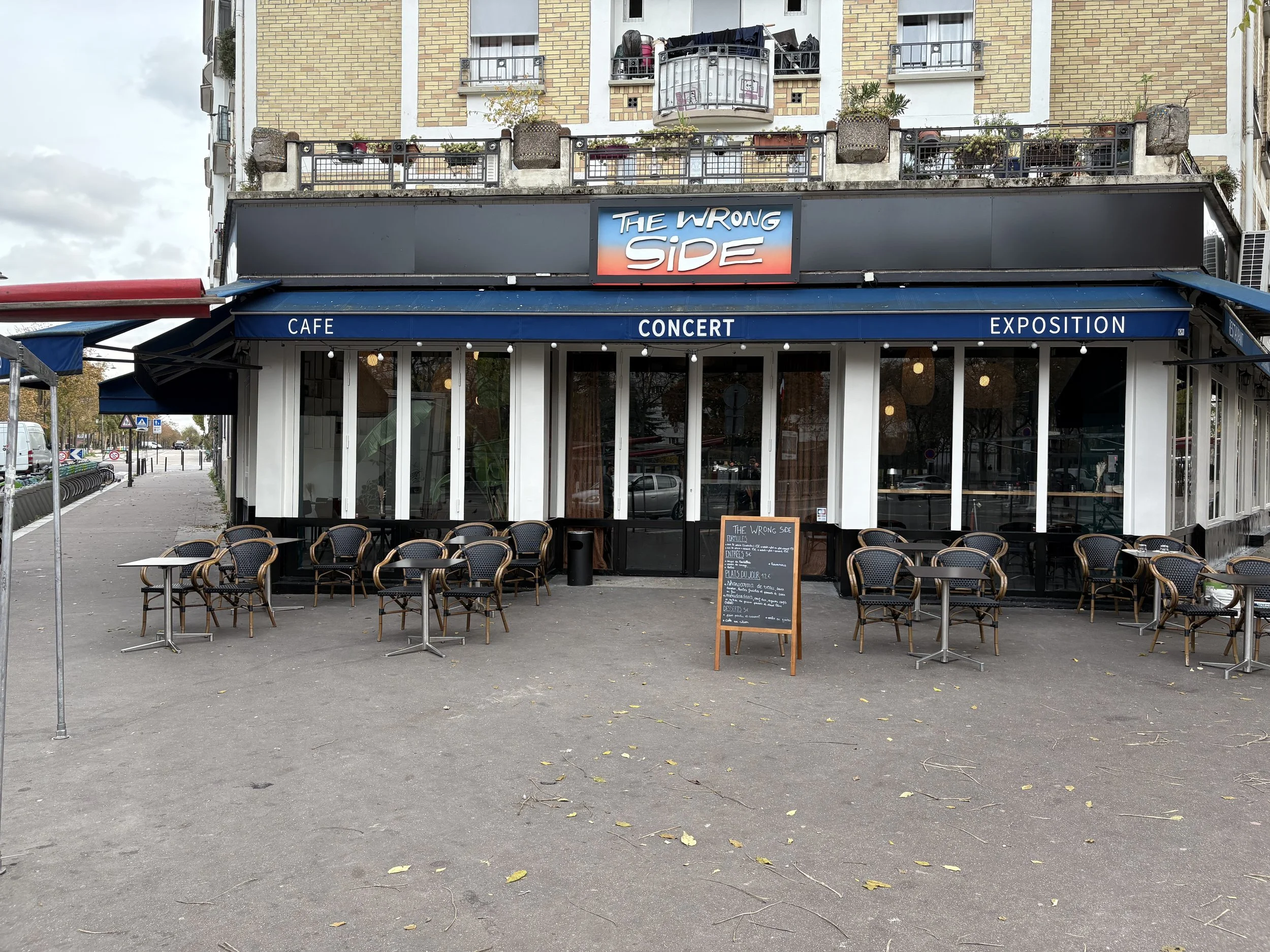 Une façade de café avec des tables et des chaises à l'extérieur. Il y a un panneau noir avec un menu écrit à la craie. La signalisation indique Café, Concert, Exposition, et un panneau lumineux affiche 'The Wrong Side'.