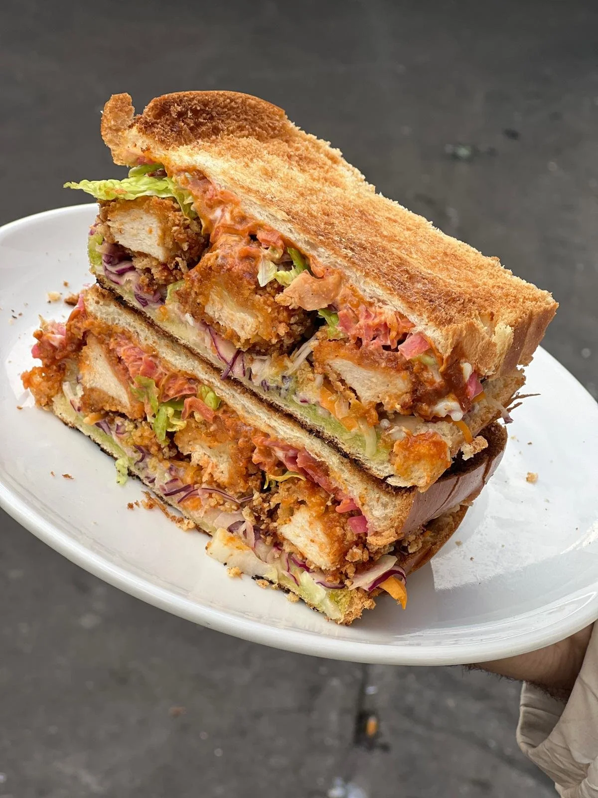 Deux sandwichs empilés avec du pain grillé, garnis de poulet frit, de laitue, de tomate, de fromage et de diverses sauces, sur une assiette blanche. En arrière-plan, une surface sombre.