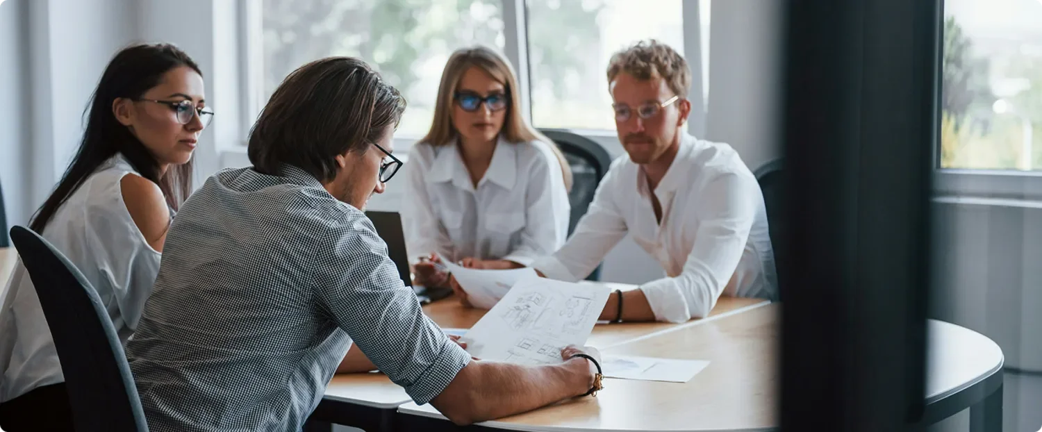 Quatre personnes en réunion dans une salle de réunion, discutant autour de documents. Deux femmes et deux hommes, portant des vêtements professionnels, assis autour d'une table.