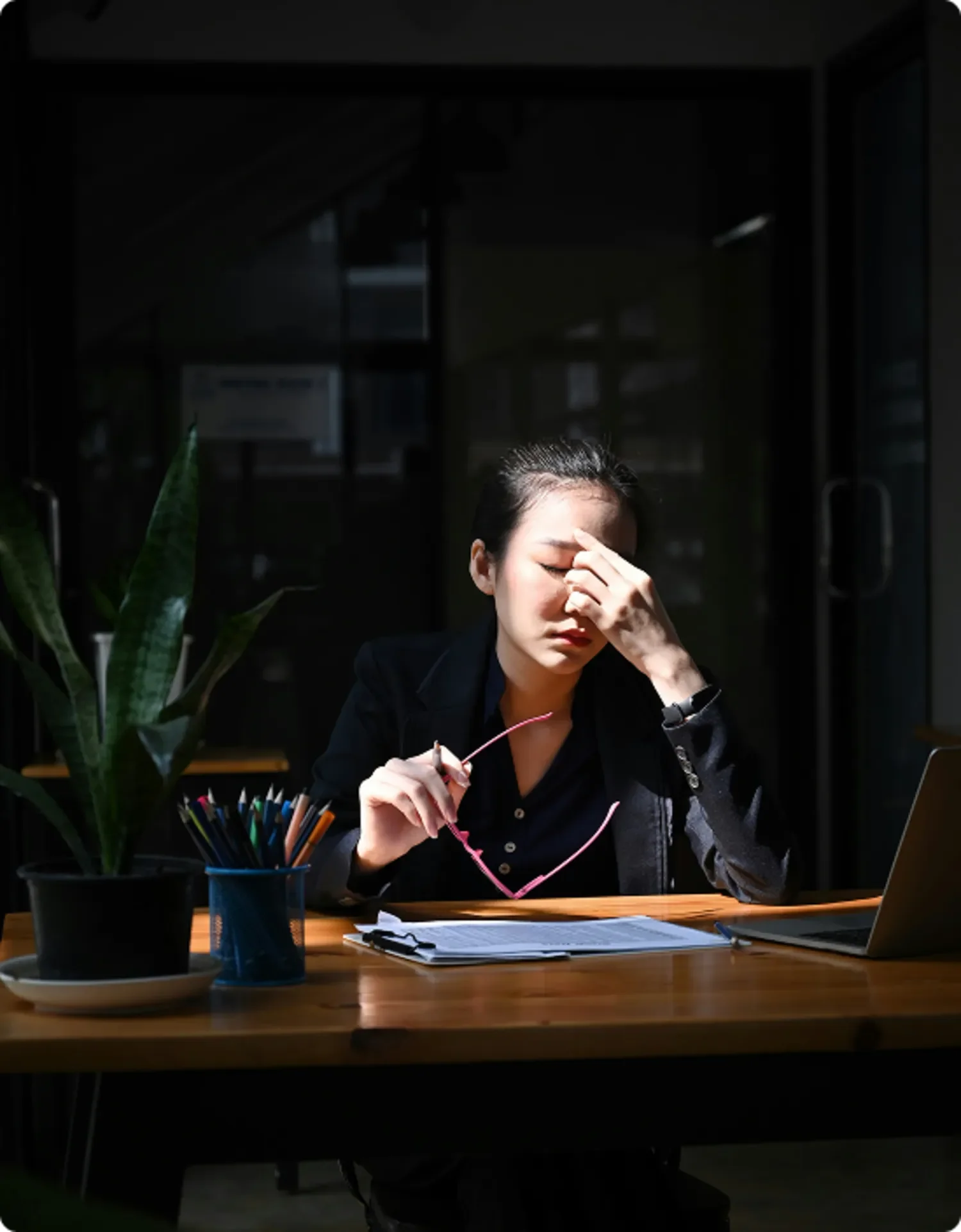 Une femme semble stressée ou fatiguée, assise à un bureau avec un ordinateur, des papers, des crayons, et une plante, dans une pièce sombre. Elle se tient la tête avec une main, l'air préoccupée.